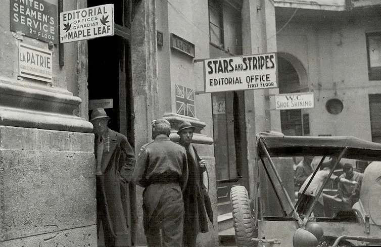 Black-and-white photo of a few men standing near a doorway, in front of signage for the Stars and Stripes editorial office.