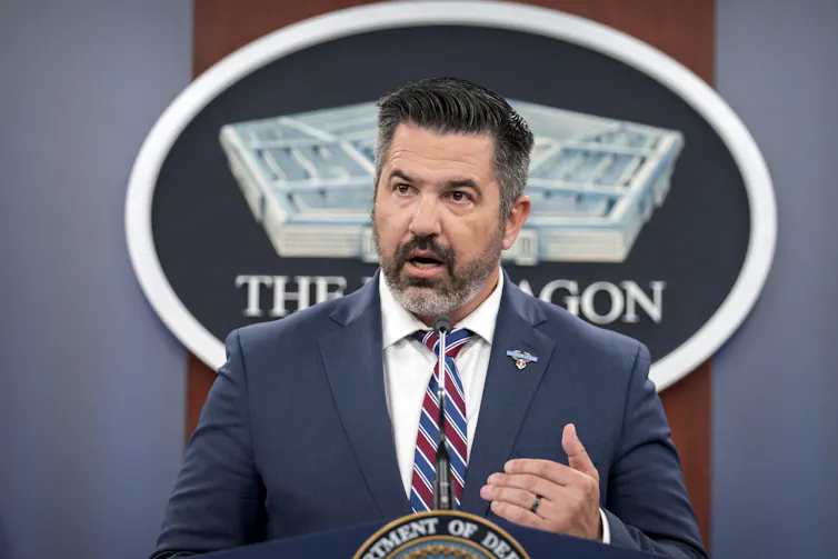 Middle-aged man with beard wearing a suit stands at a lectern and speaks into a microphone.