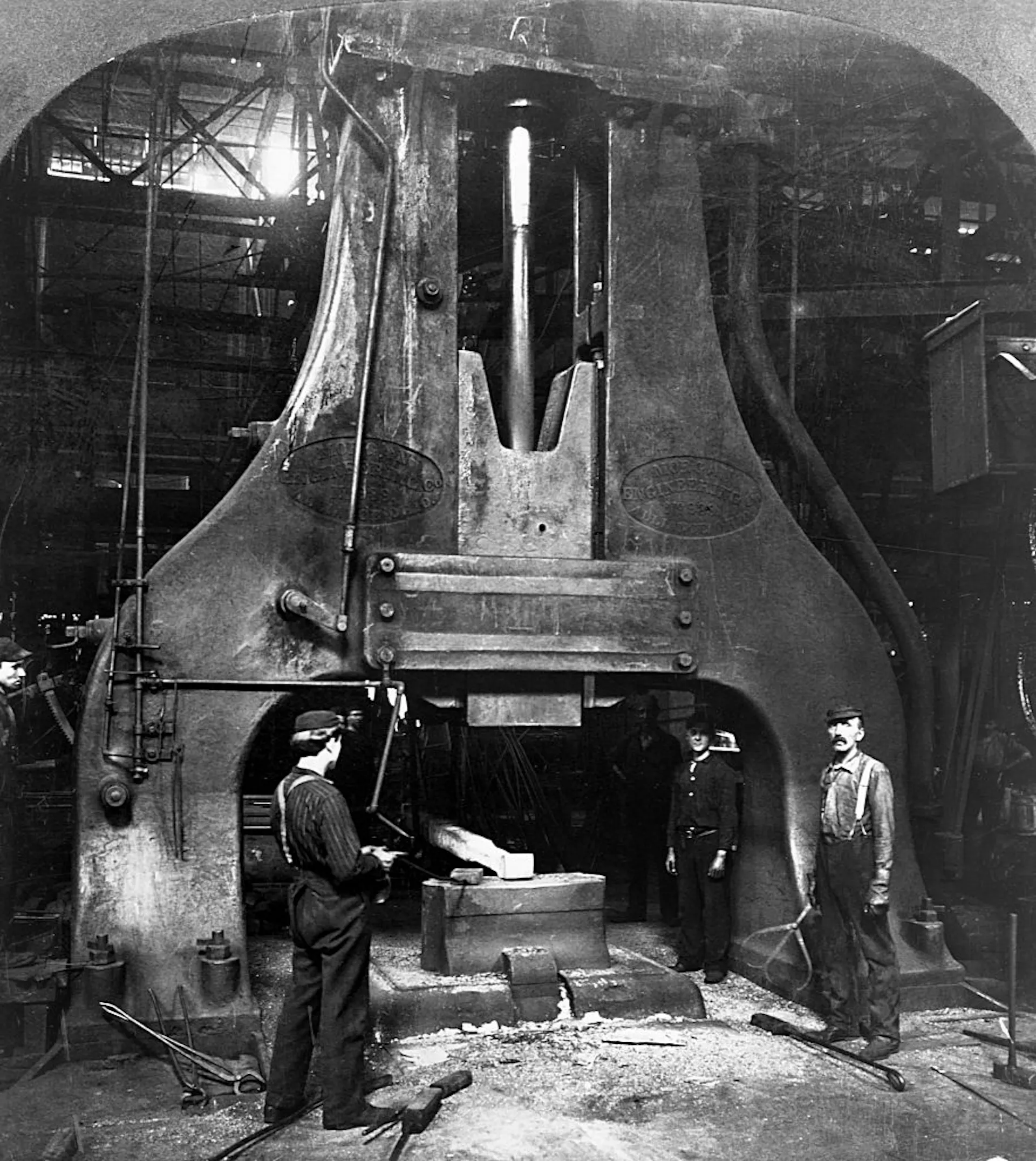 Steel workers stand next to a trip hammer in a steel mill in a black-and-white photo.