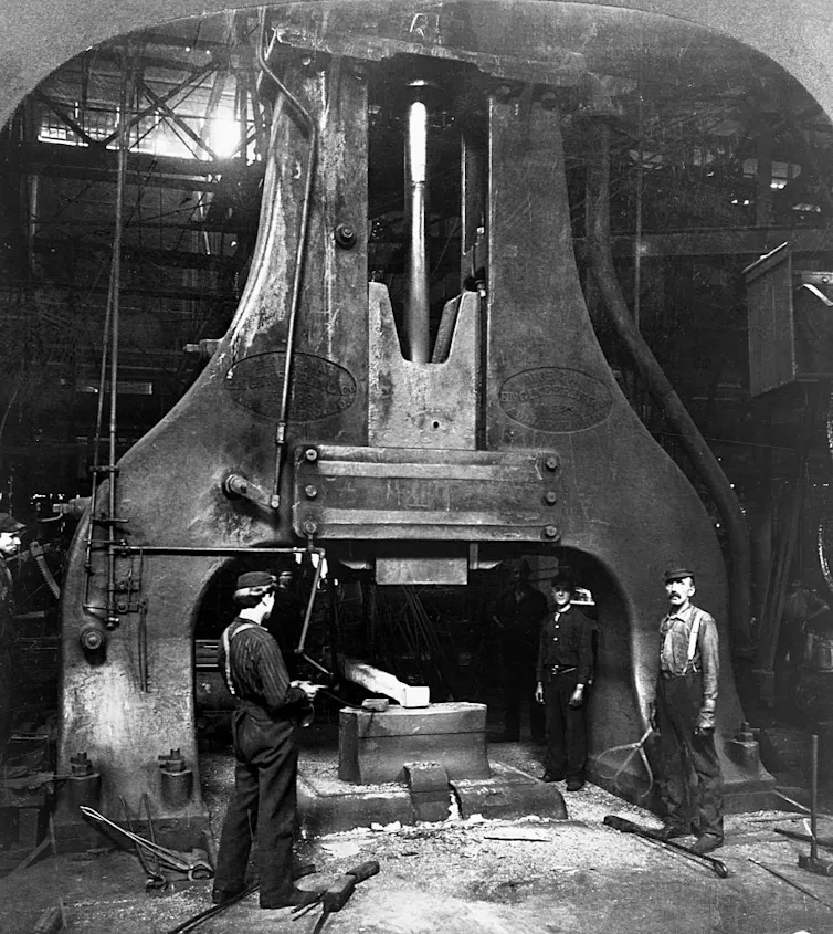 Steel workers stand next to a trip hammer in a steel mill in a black-and-white photo.