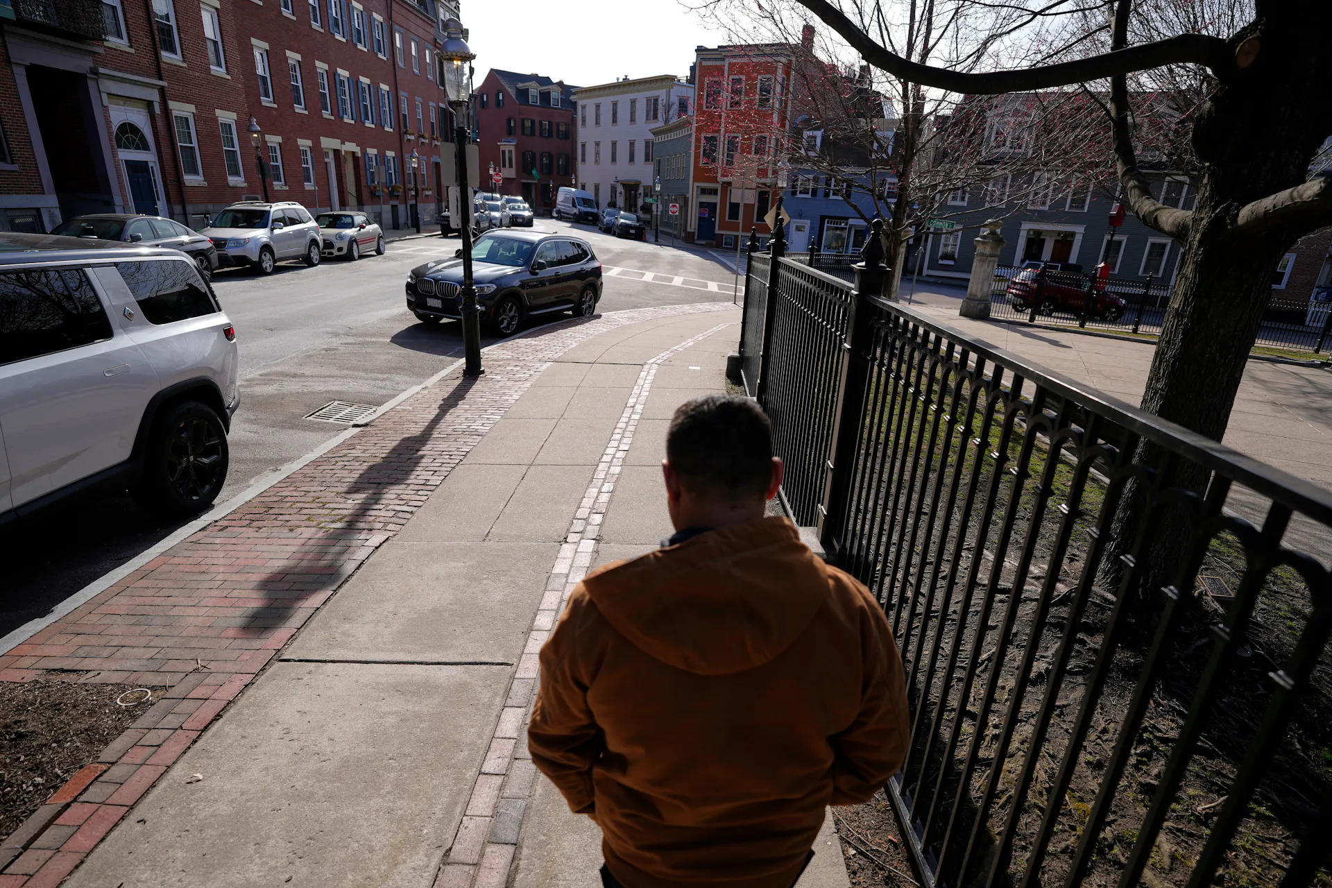 A man walks away from the camera along a sidewalk beside a fence, on a street lined with houses and parked cars.
