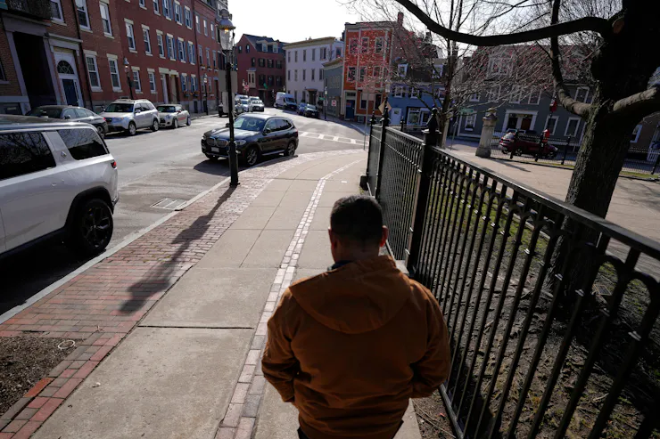 A man walks away from the camera along a sidewalk beside a fence, on a street lined with houses and parked cars.