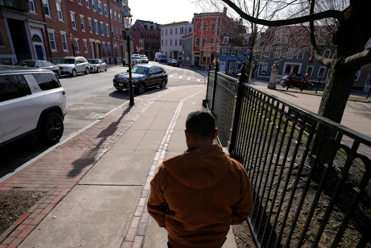 A man walks away from the camera along a sidewalk beside a fence, on a street lined with houses and parked cars.