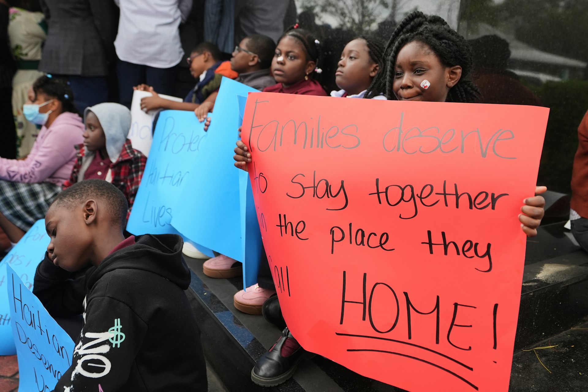 A young person at a rally holds a sign that reads 'families deserve to stay together in the place they call home.'