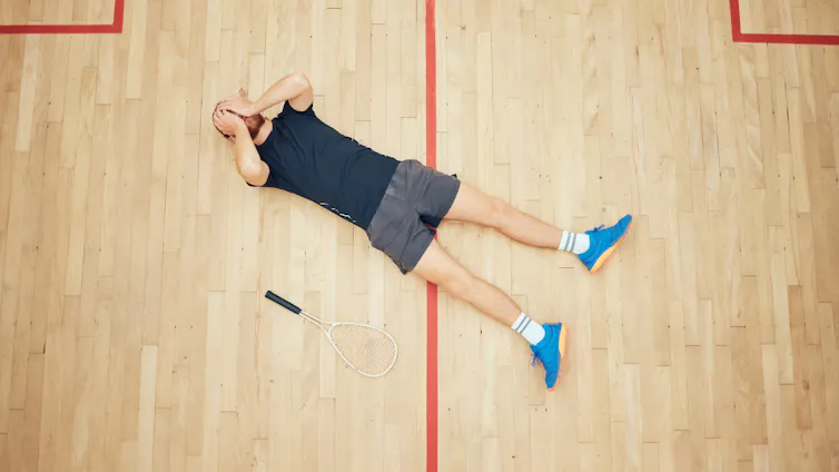 A man lays on the floor of a squash court with her hands over his eyes.