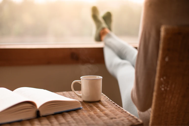 Person sitting in chair, with feet up on windowsill, resting after reading a book. An open book and mug are on the side table next to the chair.