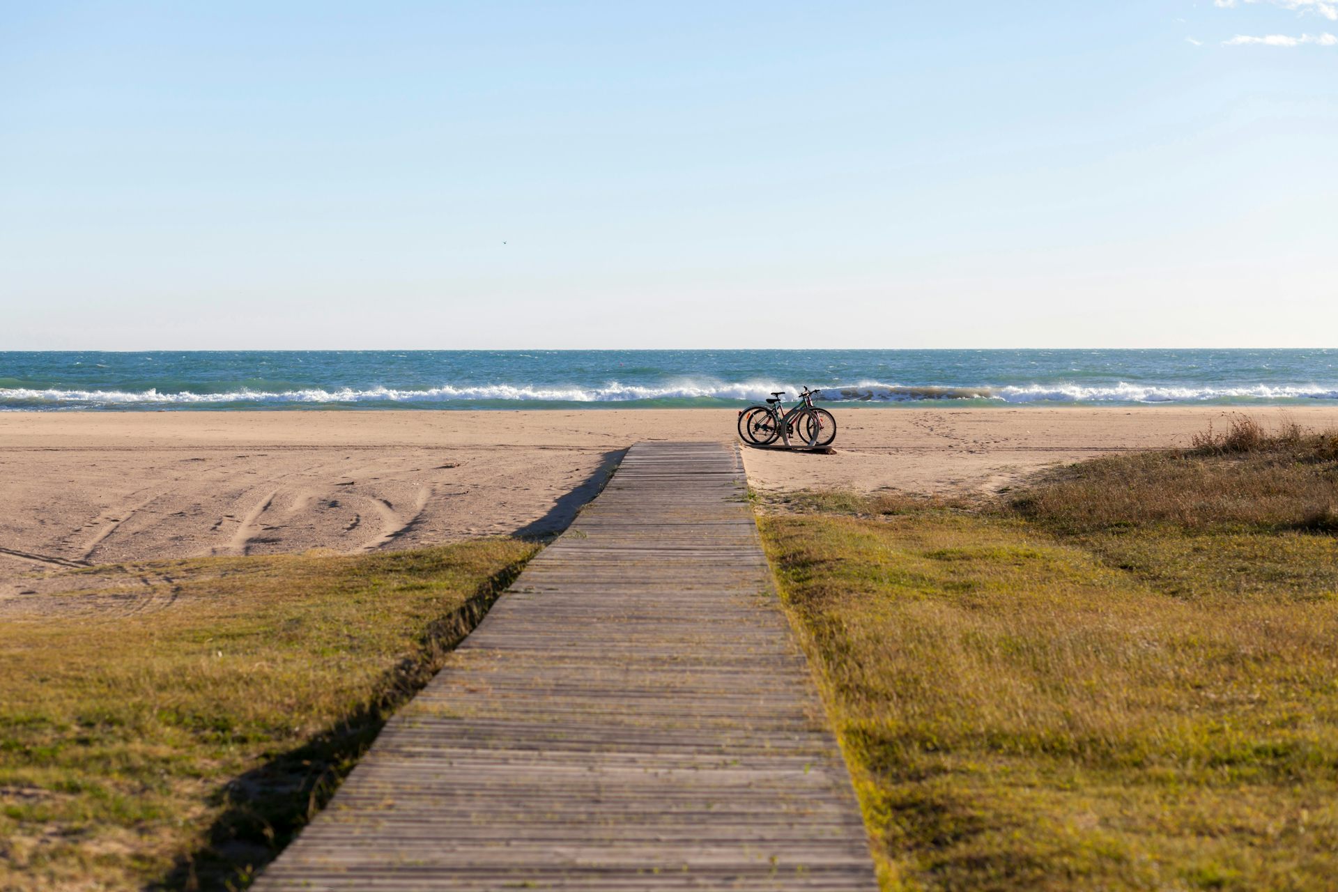 Un passeig de fusta porta a una platja de sorra. A la dreta del passeig hi ha dos bicicletes.