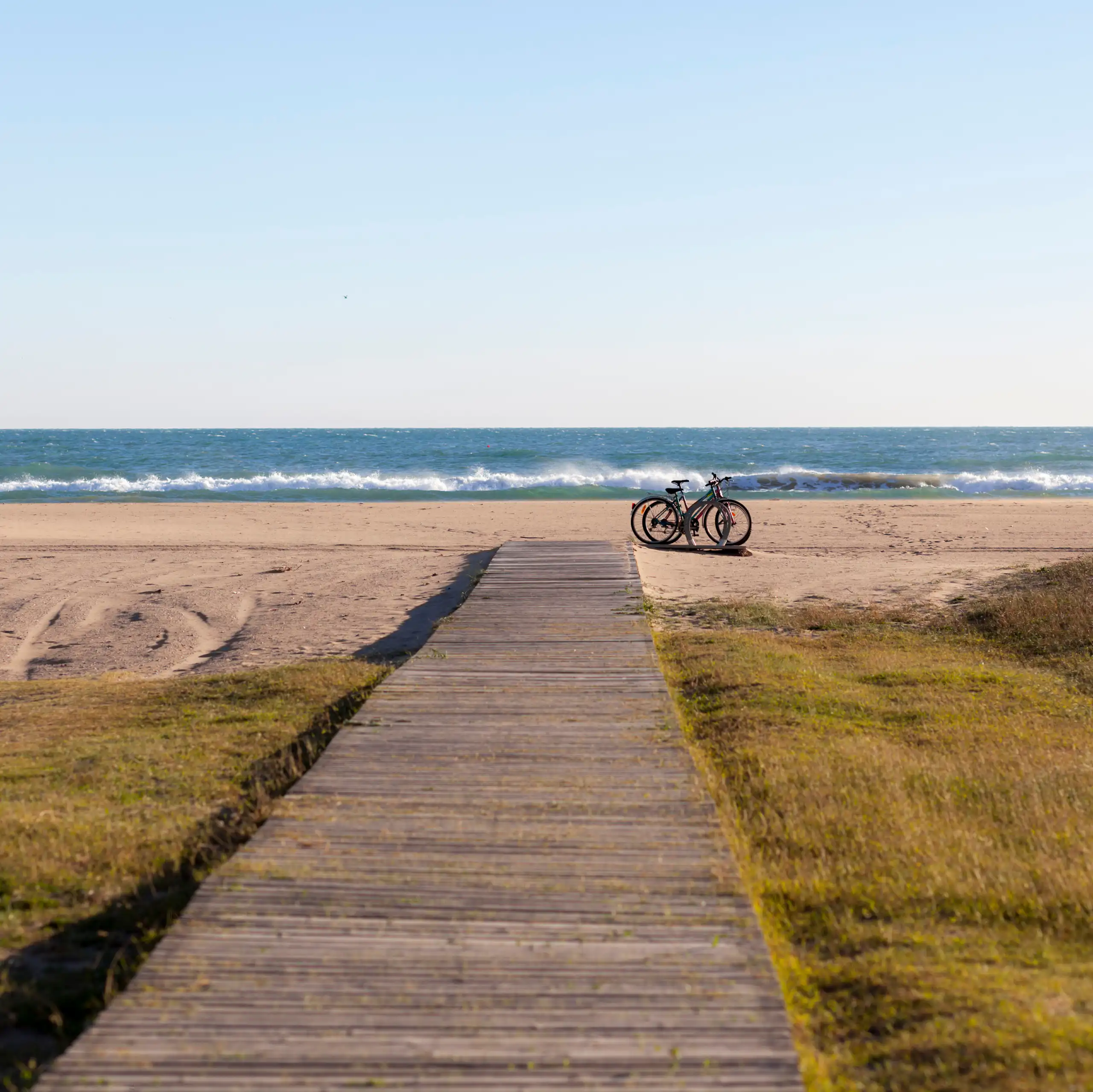 Un passeig de fusta porta a una platja de sorra. A la dreta del passeig hi ha dos bicicletes.