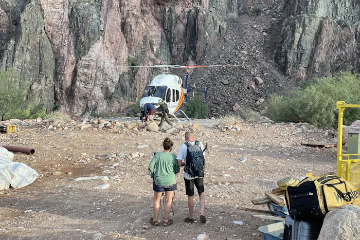 Two people wait on a rocky canyon floor for a helicopter in the distance.