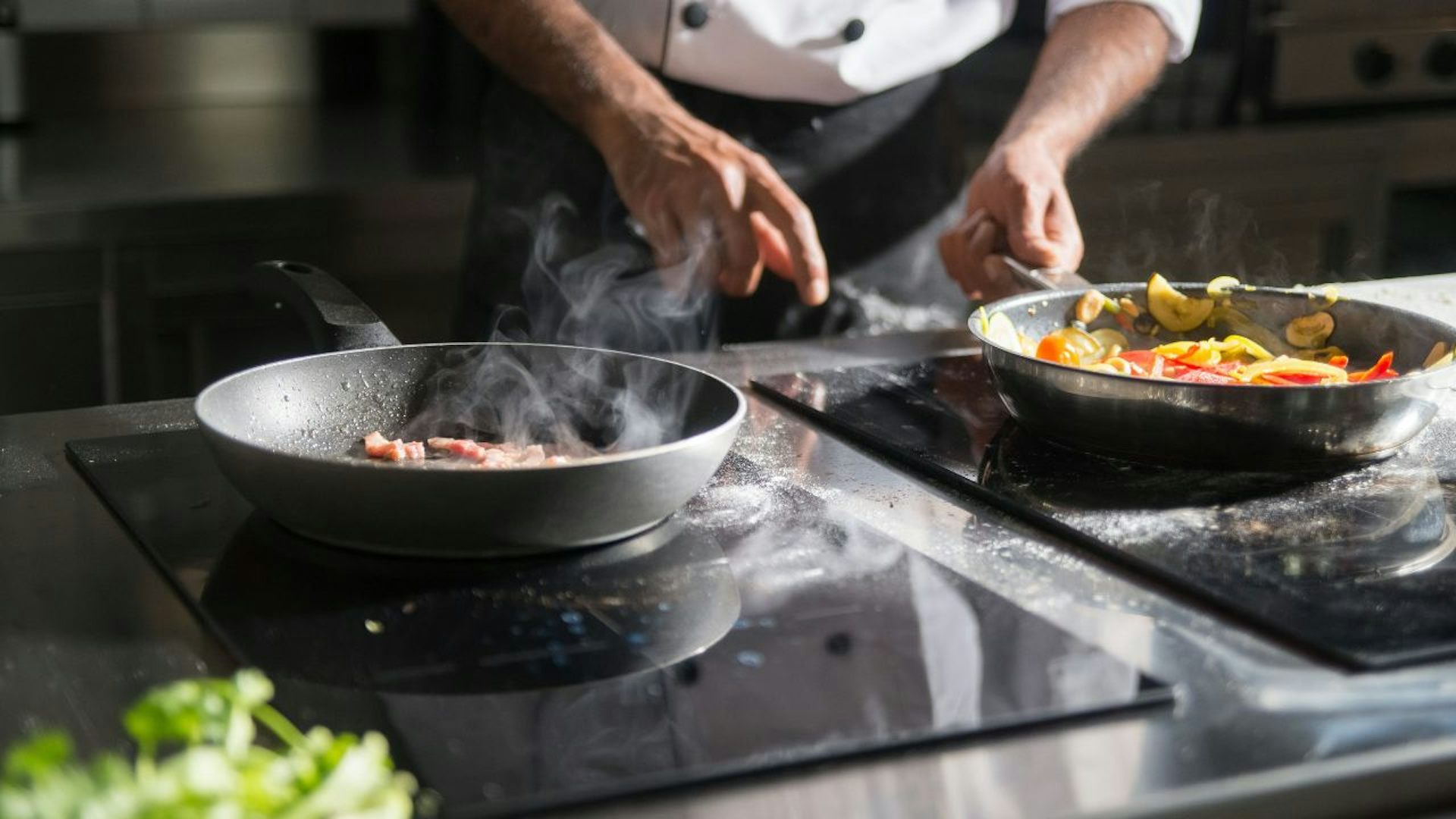 hands are seen handling a pan with food on an electric stove, with a second stove with food in the foreground