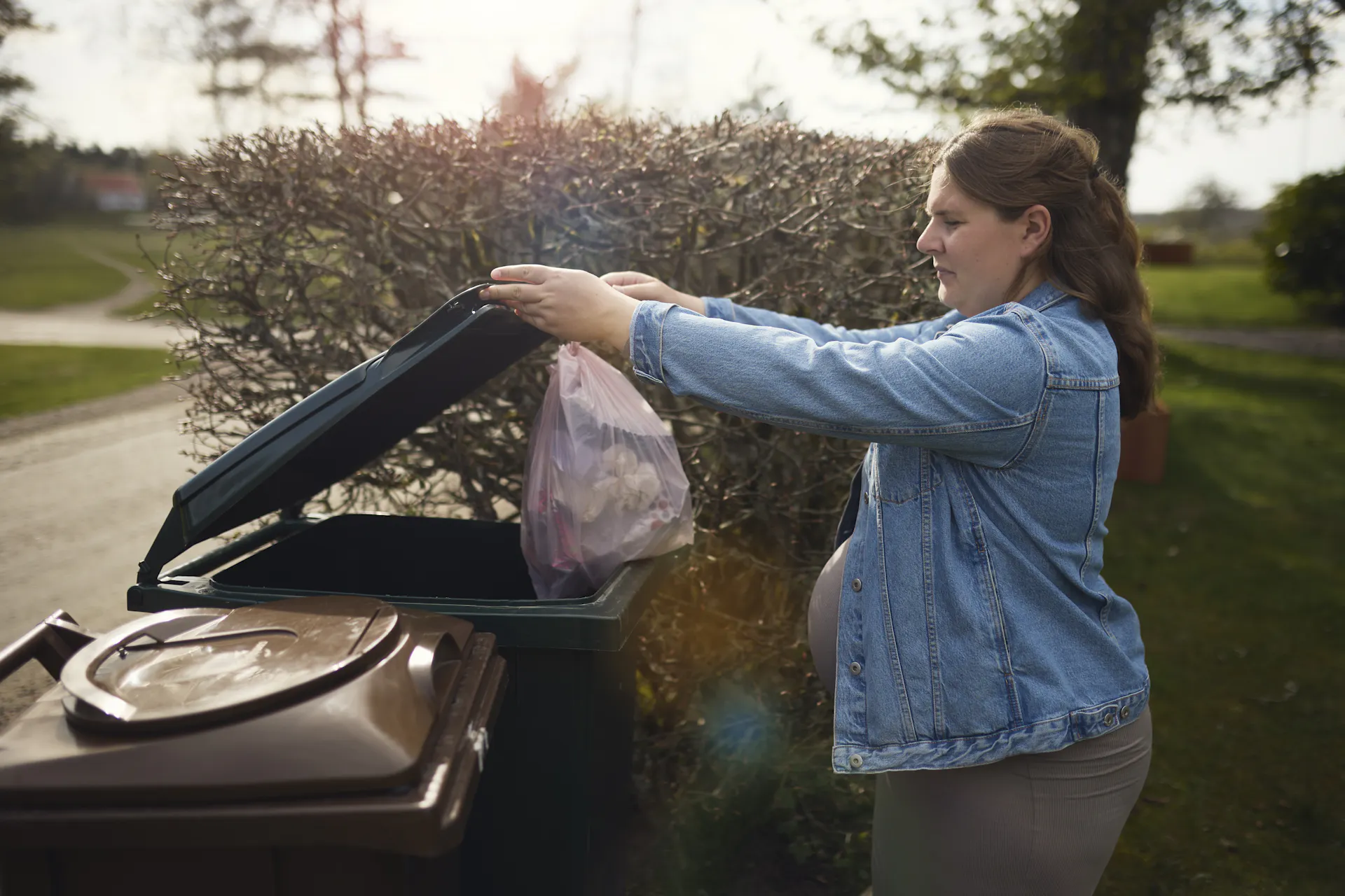 A pregnant woman throws a garbage bag into a trash can in a backyard.