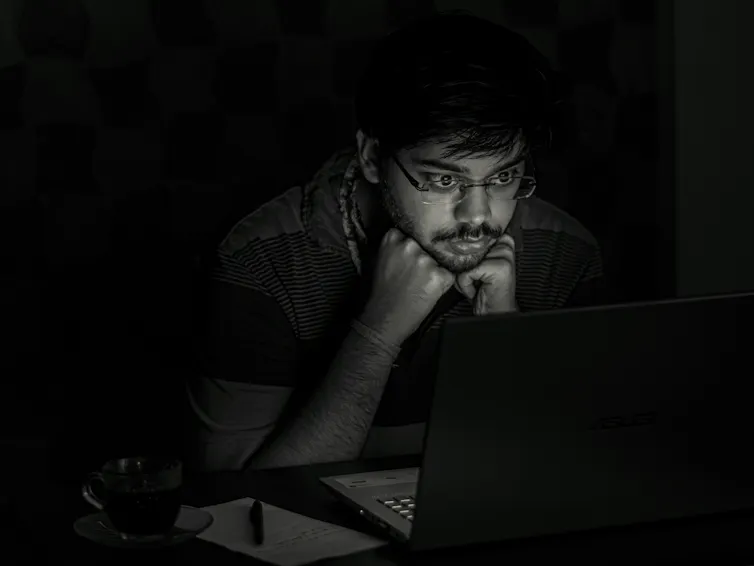Black and white photo of a man in dark room, his face illuminated by the glow of a computer screen