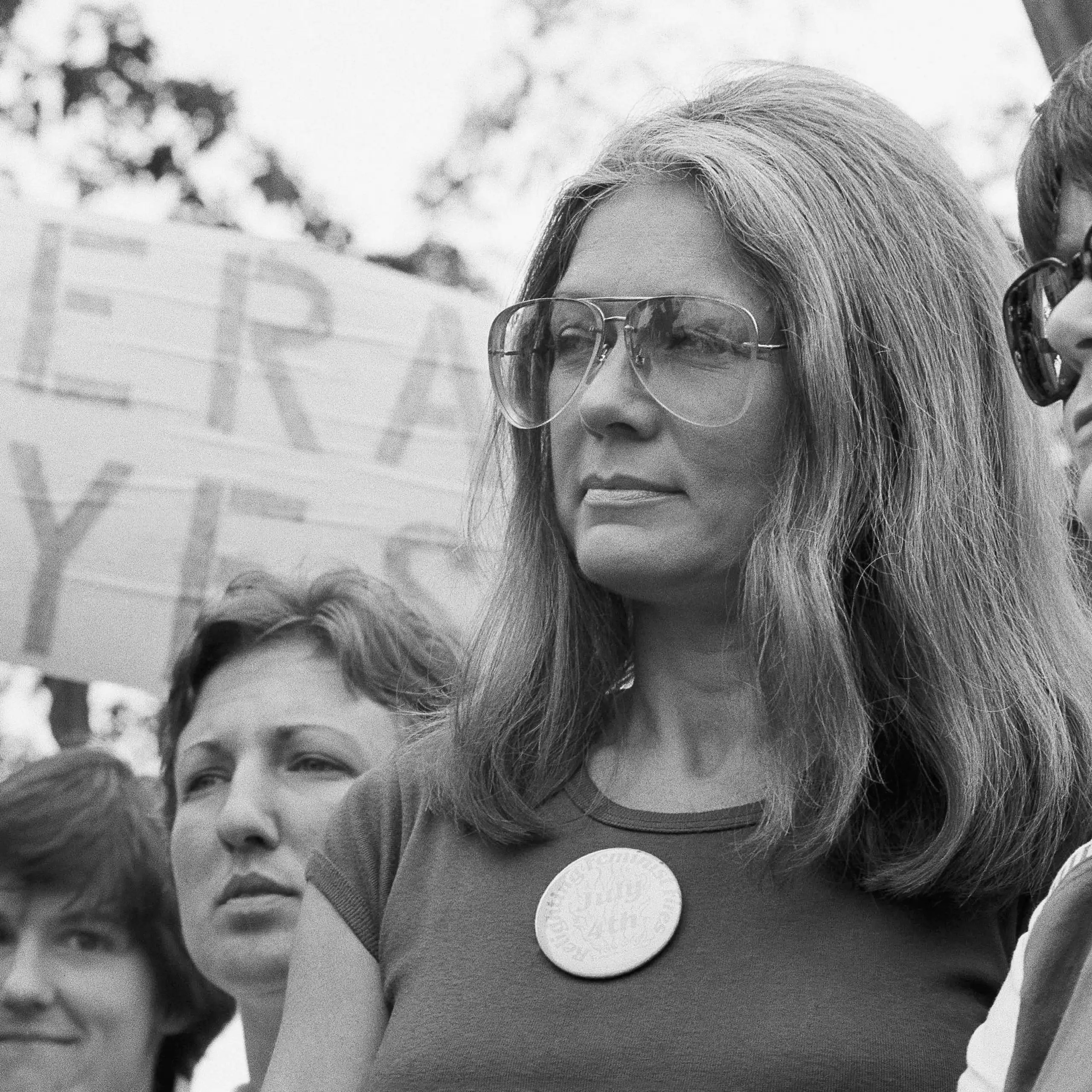 A woman in aviator sunglasses with a discerning look on her face in front of a sign that says ERA YES.