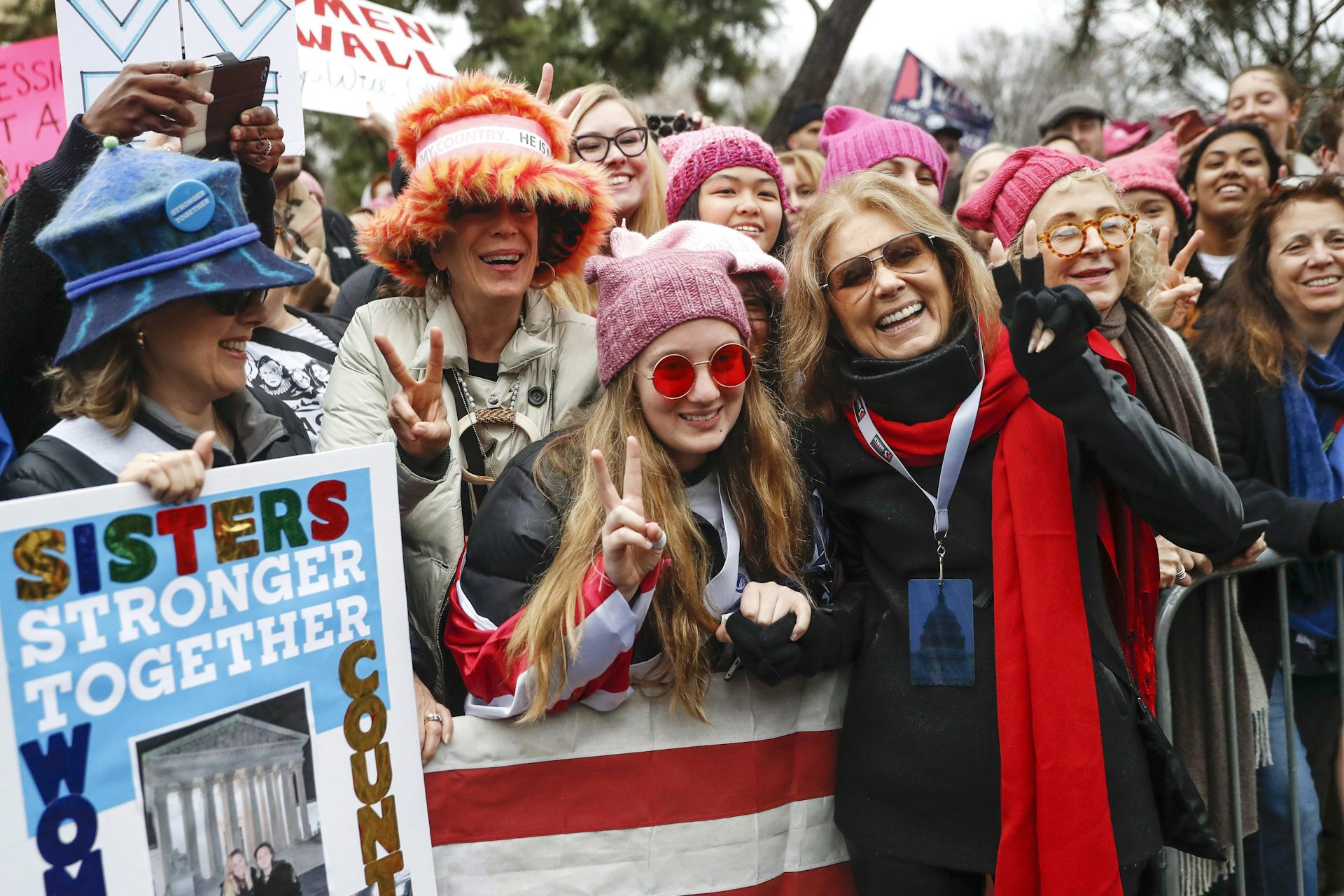 People seen standing in pink hats with placards, one that says sisters stronger together, at a rally.
