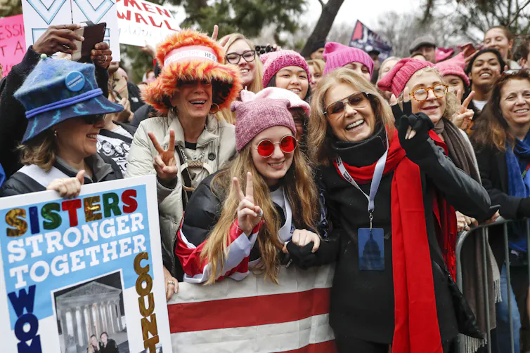 People seen standing in pink hats with placards, one that says sisters stronger together, at a rally.