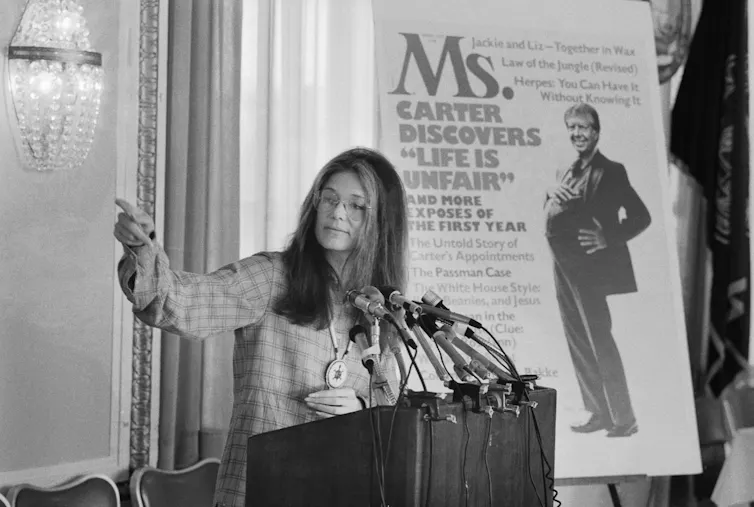 A woman stands at a lectern in front of a large magazine cover and speaks.