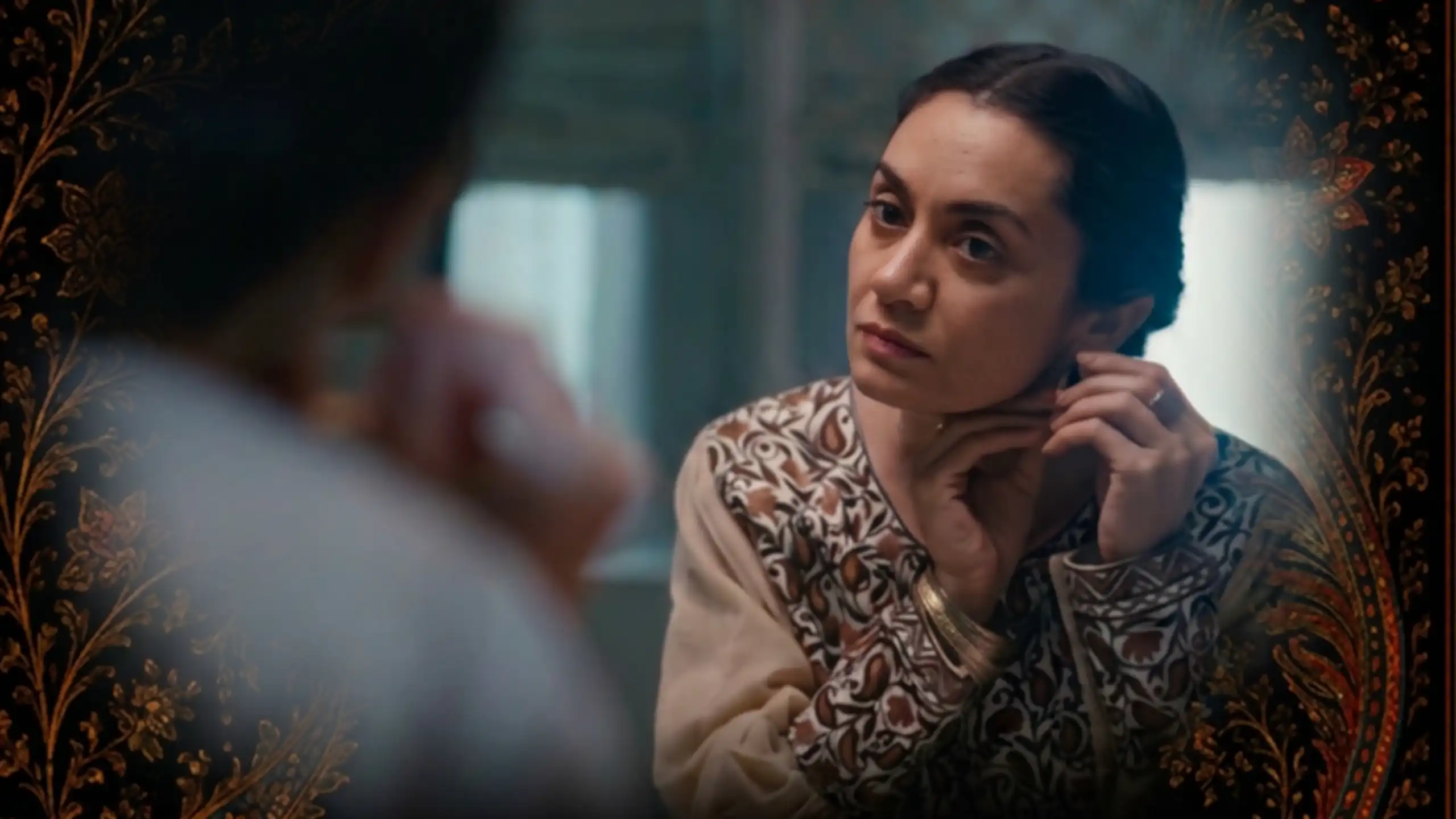 A brown woman will her hair pulled back looks into the mirror as she changes her earring.
