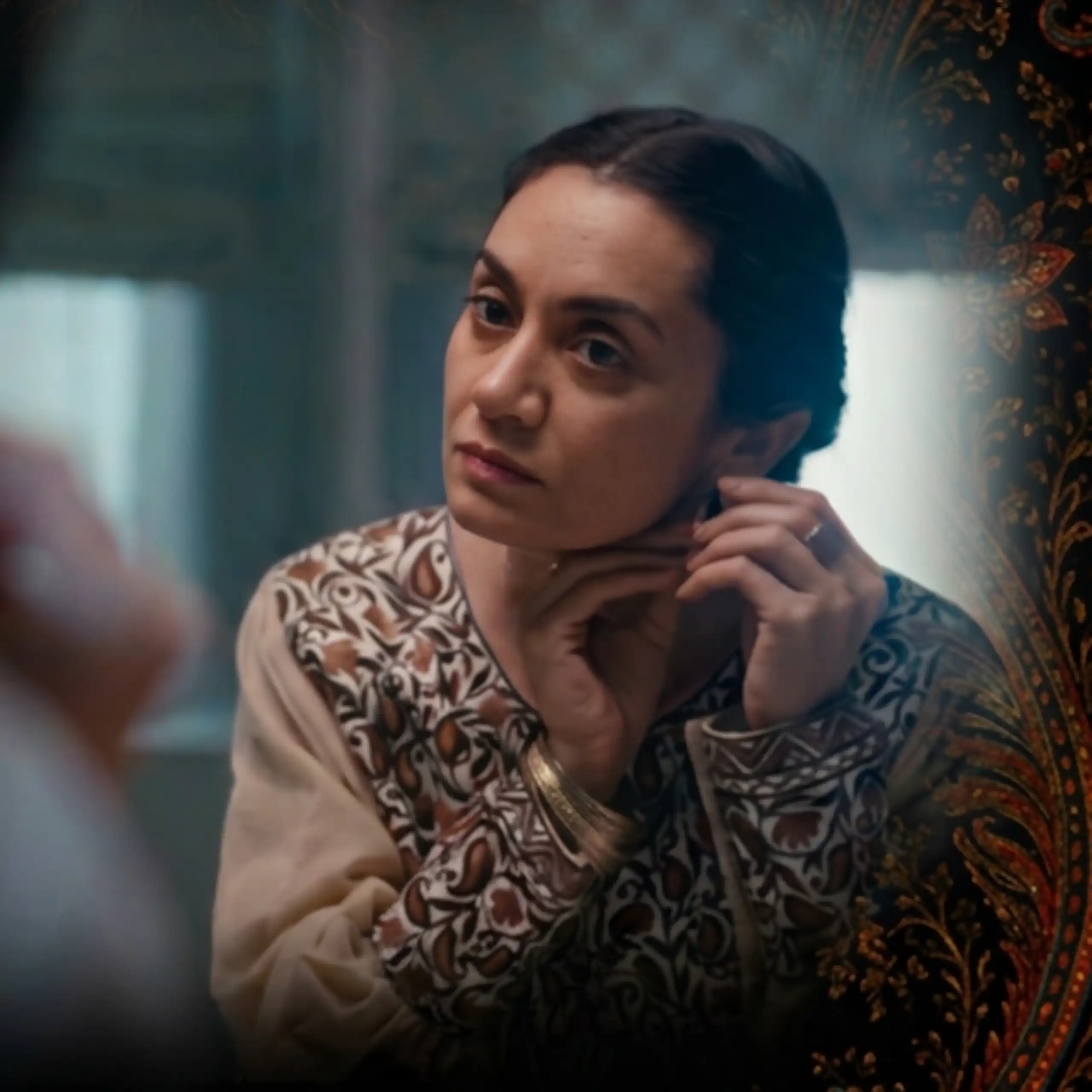 A brown woman will her hair pulled back looks into the mirror as she changes her earring.