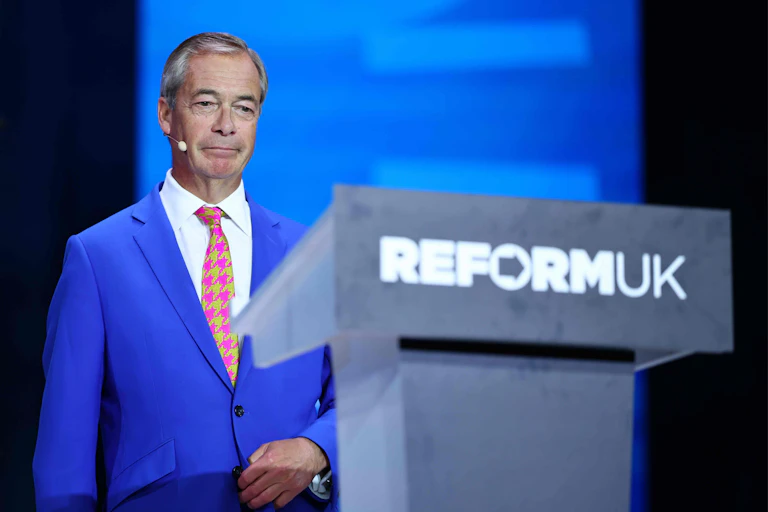 nigel farage approaching a lectern with Reform UK on it against a blue backdrop.