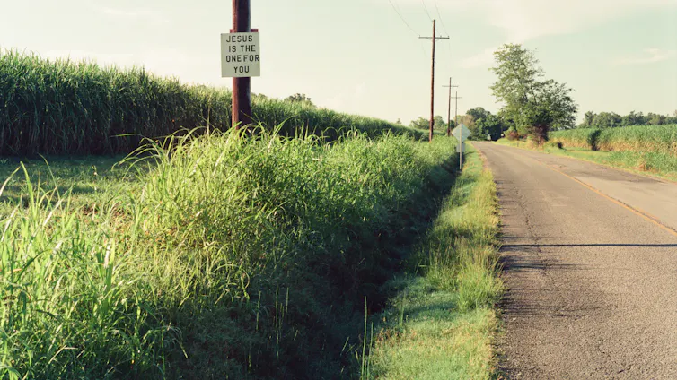 A rural road runs alongside a field of tall crops, with a small sign that says 'Jesus is the one for you' hanging from an electrical pole.