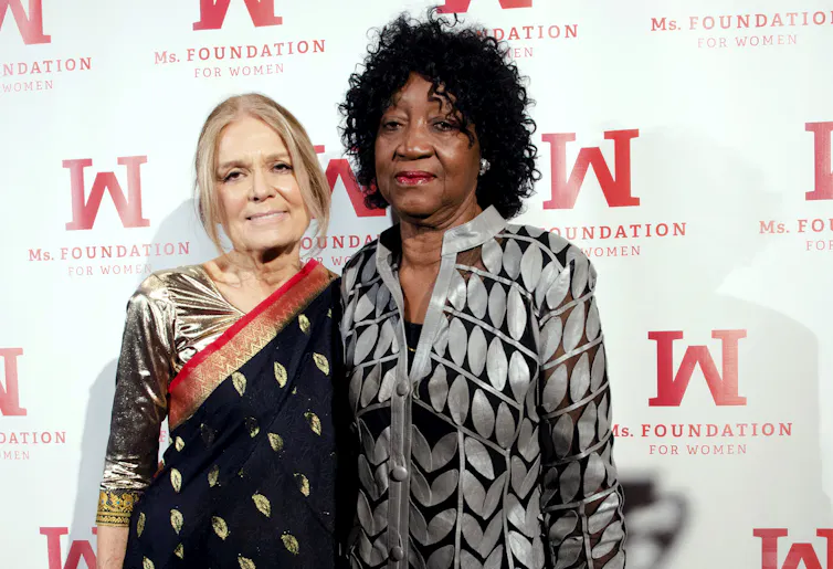Two women, one white and one Black, pose in front of a white backdrop with their arms around each other.