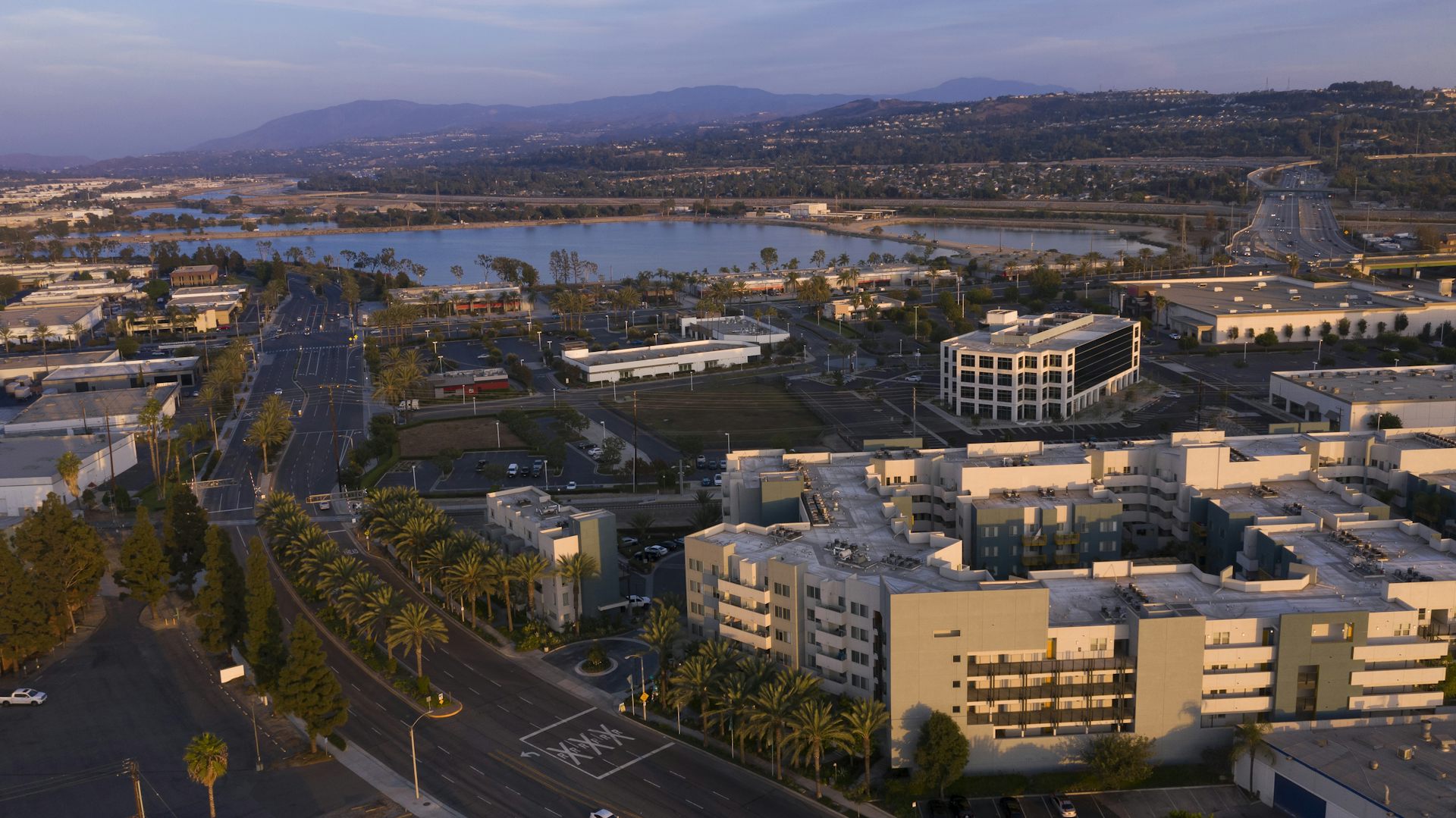 An aerial photo with palm trees, buildings, water and mountains at sunset.