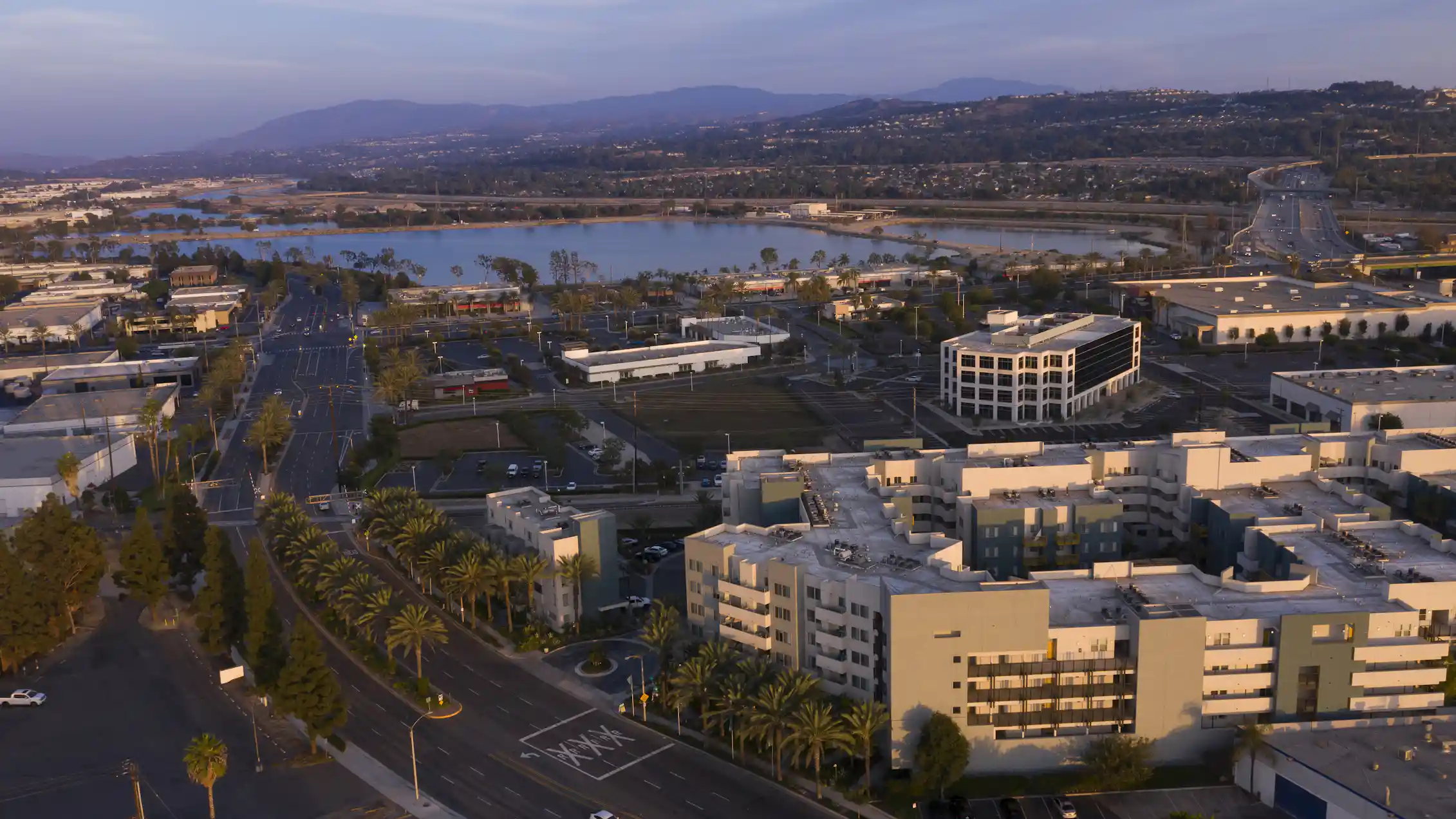 An aerial photo with palm trees, buildings, water and mountains at sunset.