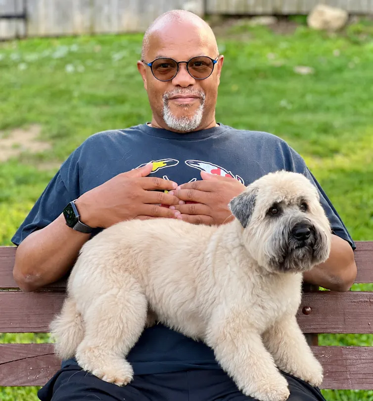 A man in sunglasses sits on park bench with small white dog standing on his lap