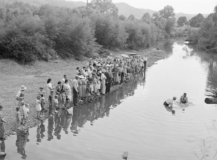 A black-and-white photo of a row of people on the shore of a creek, watching as two men dunk someone in the water.
