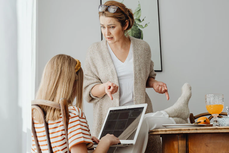 A chlid has their feet on the table while looking at a laptop and a parent is pointing at the feet.