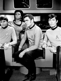A black-and-white photo of four characters wearing Starfleet uniforms, including two white men, an Asian man and a Black woman.