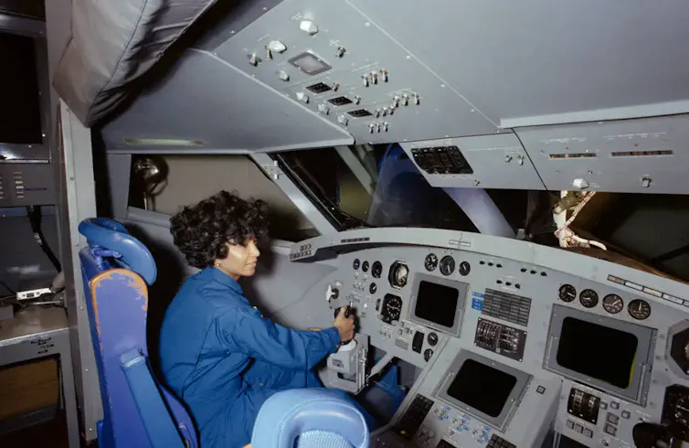 A woman wearing a blue jumpsuit sitting in the pilot's chair in a space shuttle cockpit simulator