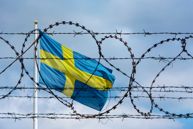 Image of the Swedish flag flying behind a barbed wire fence