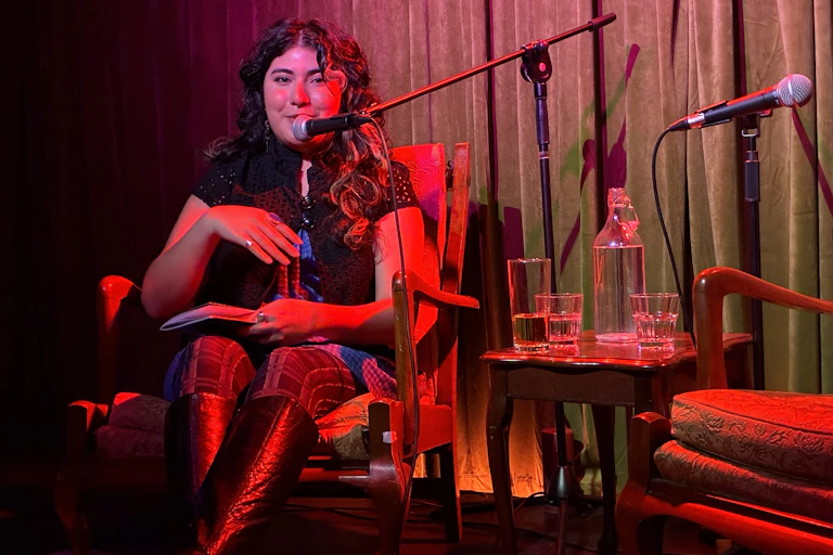 A young woman high knee-high boots and dark curls in front of a microphone, seated on stage.