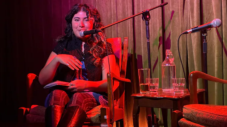 A young woman high knee-high boots and dark curls in front of a microphone, seated on stage.