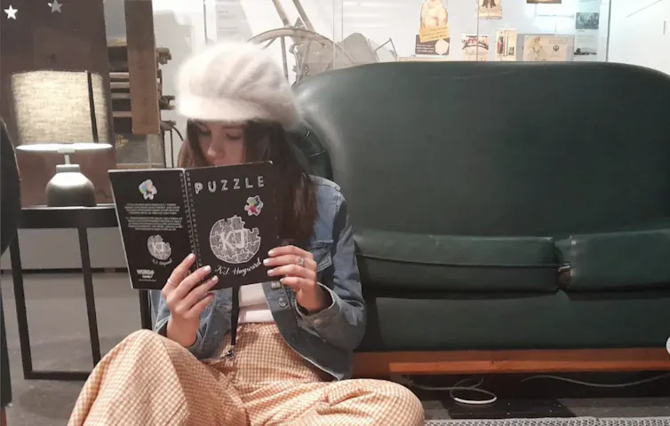 A young woman reading a book in front of a lounge, wearing a white peaked cap.