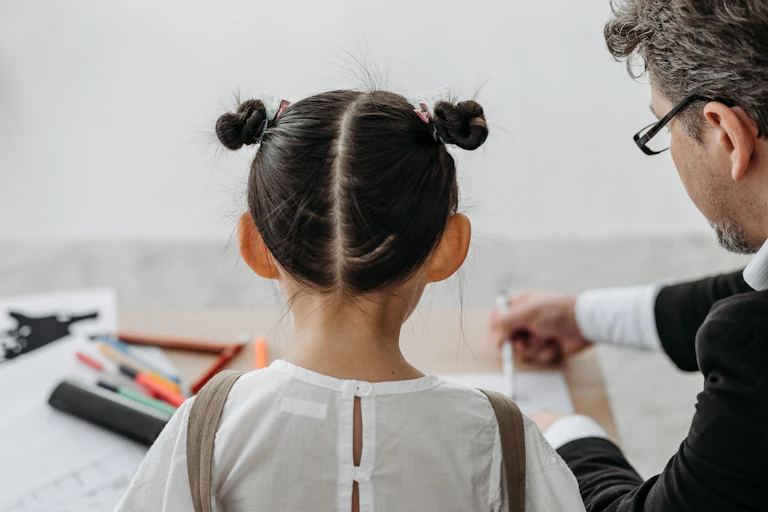 A young child and a man sit at a desk. They are working on a sheet with pens and highlighters.