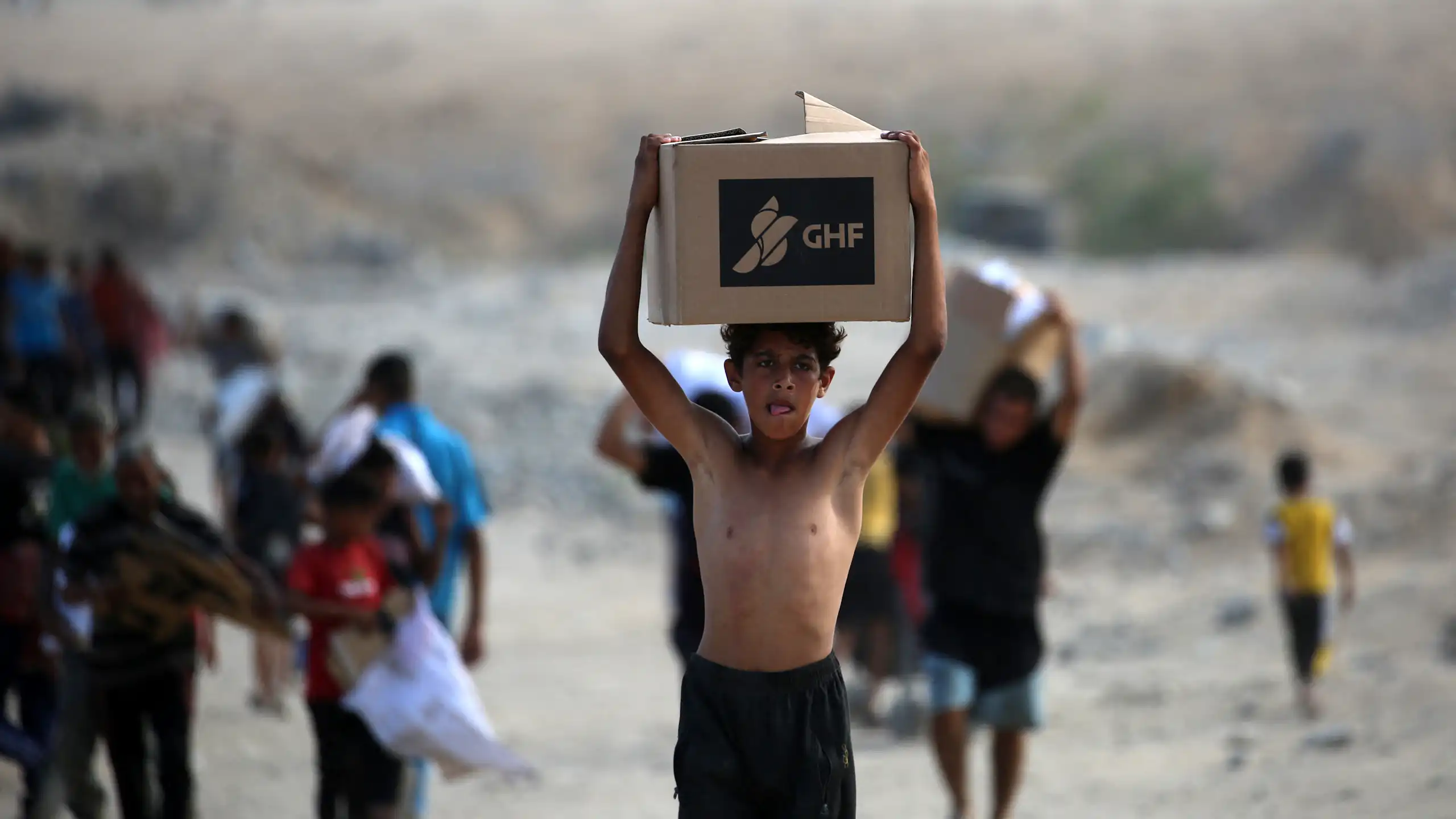 A boy carries a box saying GHF on his head as he walks through rubble