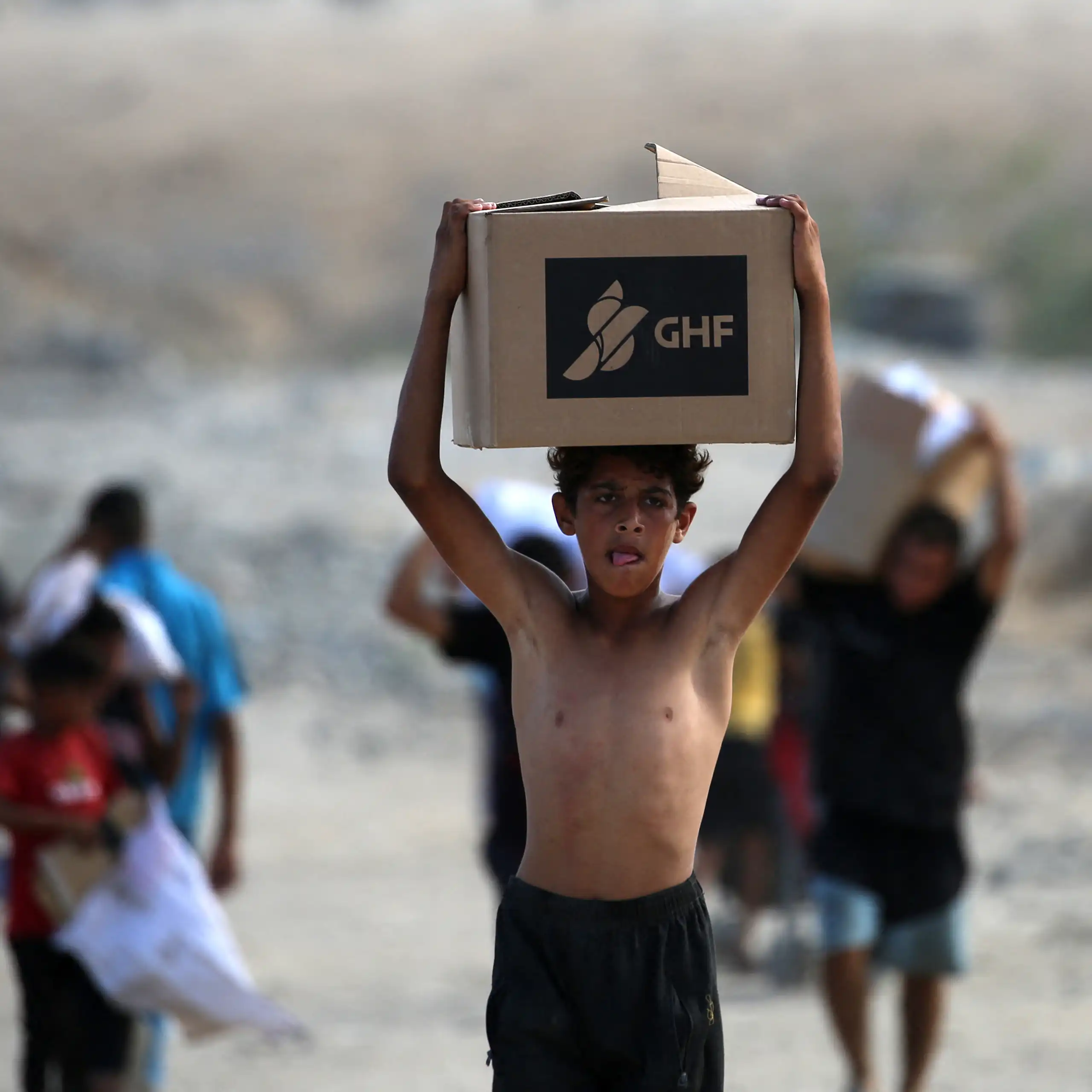 A boy carries a box saying GHF on his head as he walks through rubble