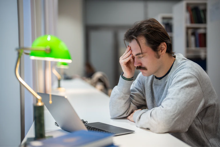 A stressed male uni student holds his hand to his head in a campus library.