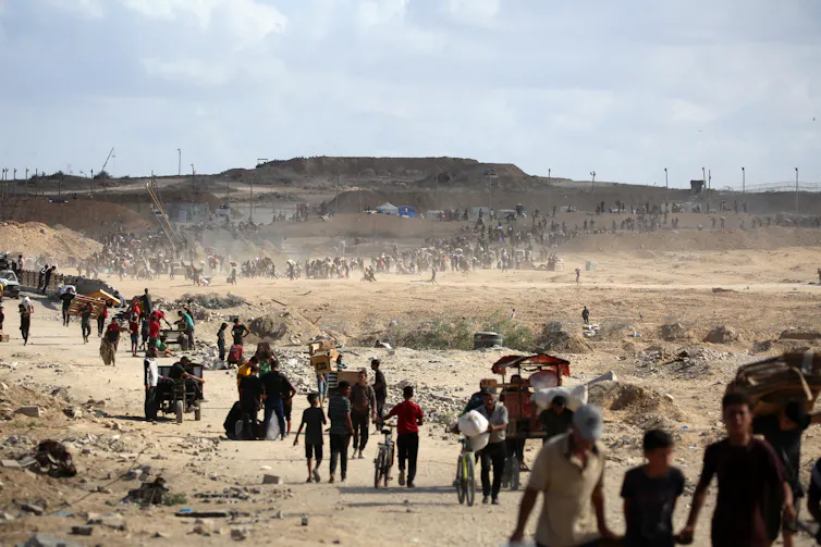 A long line of people with empty cardboard boxes and plastic bags walk through an arid landscape.