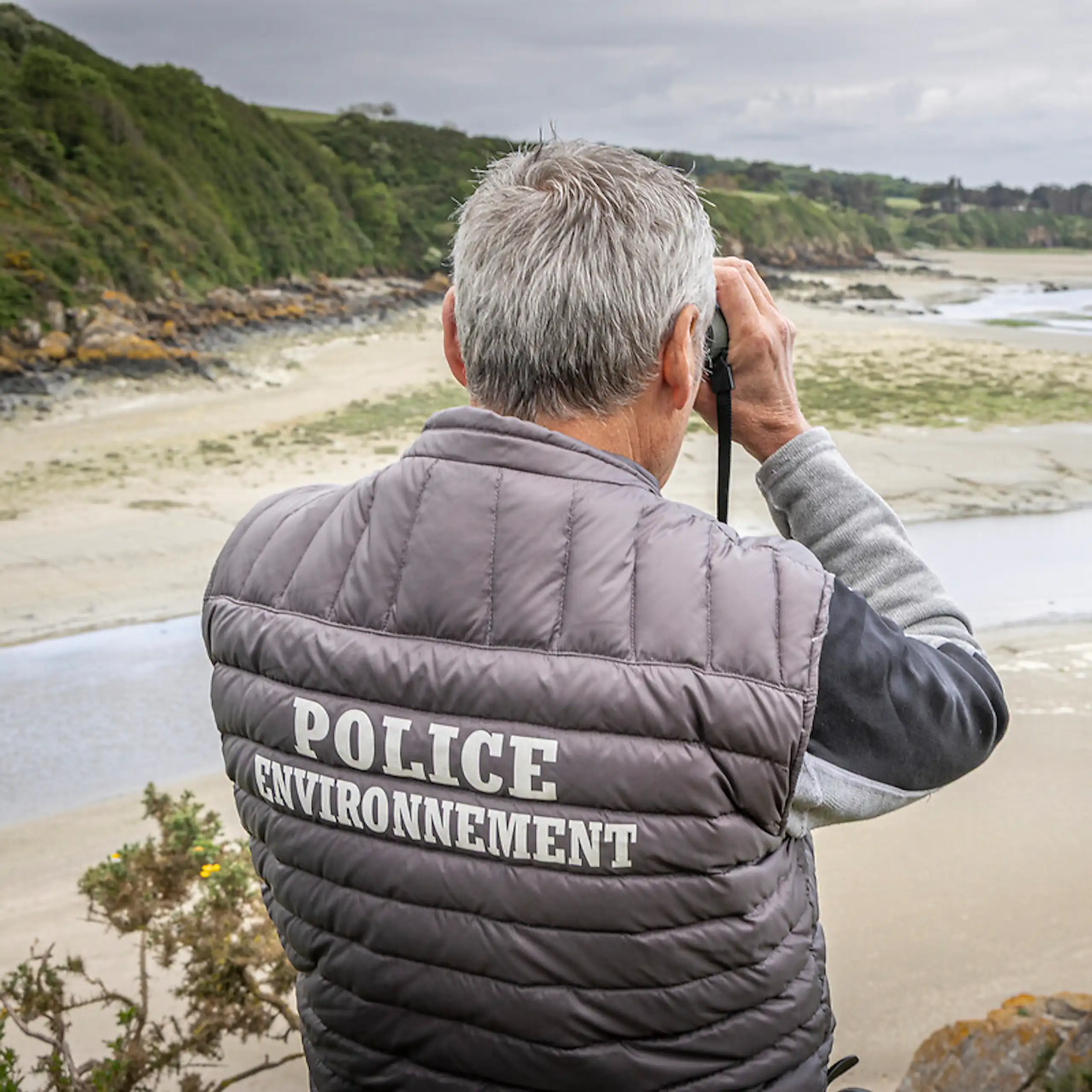 Agent de l'OFB scrutant l'horizon aux jumelles, vu de dos.