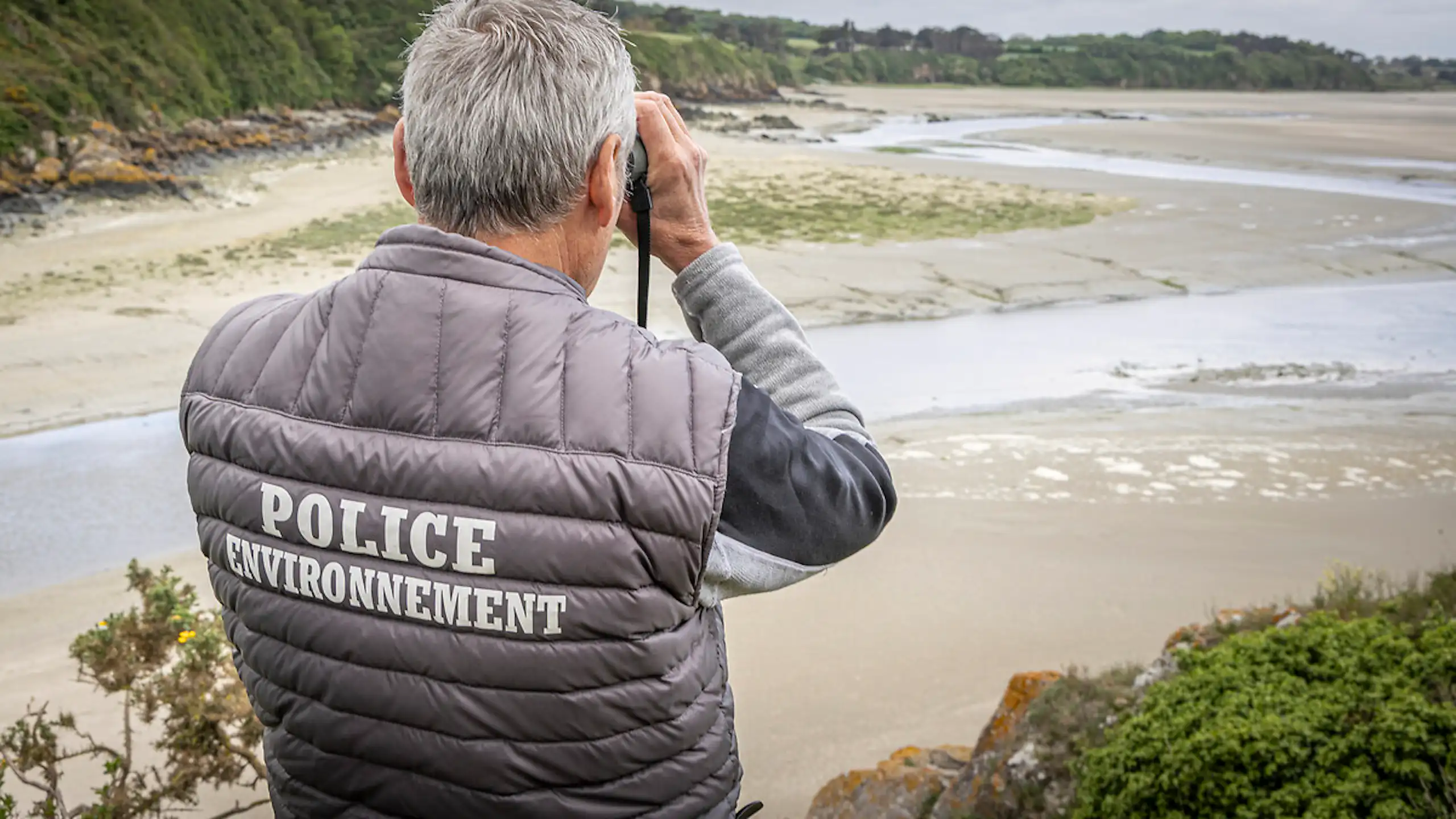 Agent de l'OFB scrutant l'horizon aux jumelles, vu de dos.