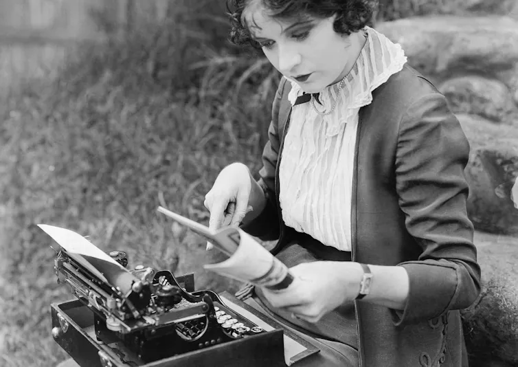 Fotografía en blanco y negro de una mujer escribiendo en una máquina de escribir mientras lee un libro.