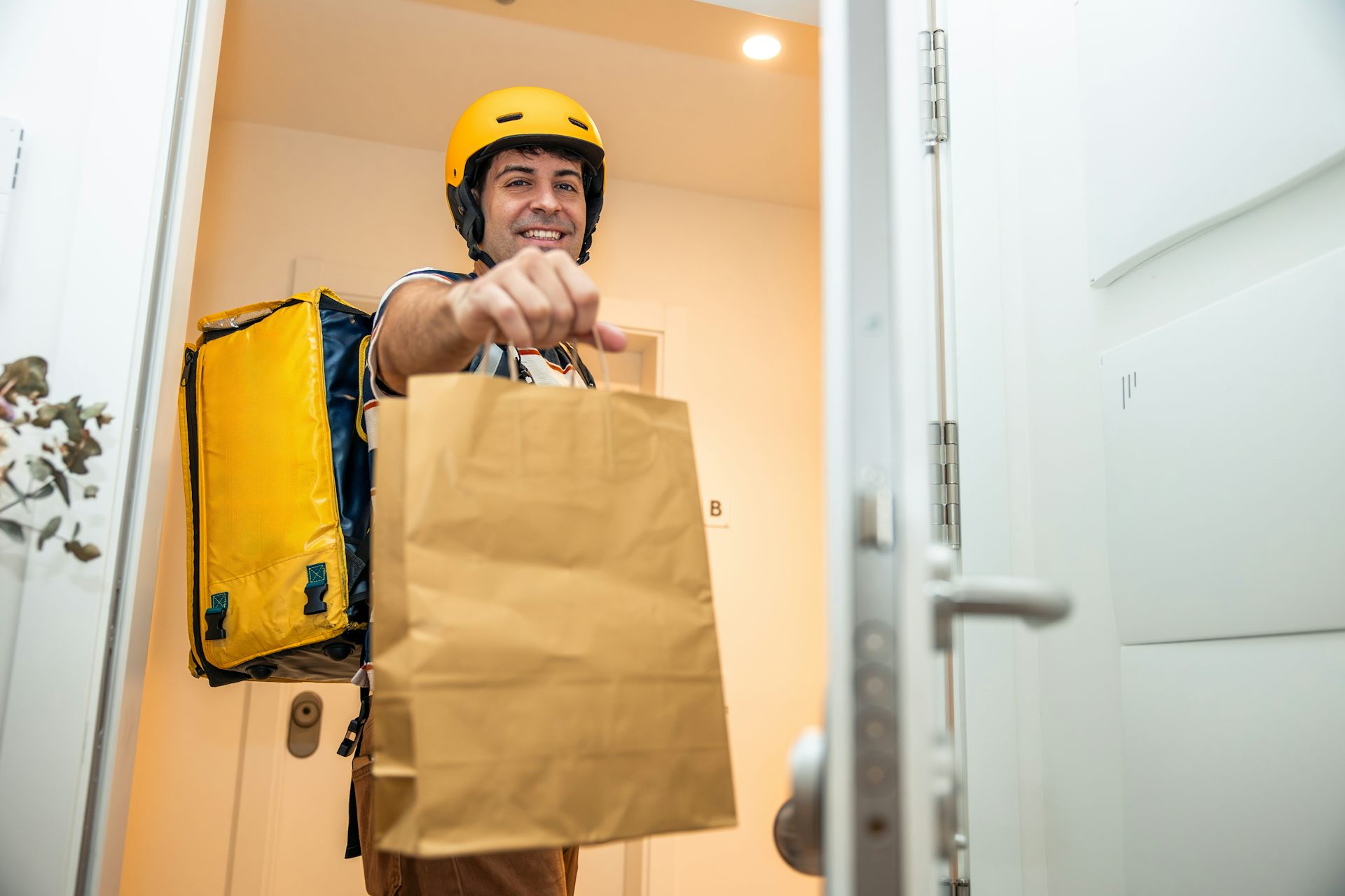 Man wearing a yellow helmet and carrying a thermal backpack delivering a takeout food order in a brown paper bag, standing at an open home door