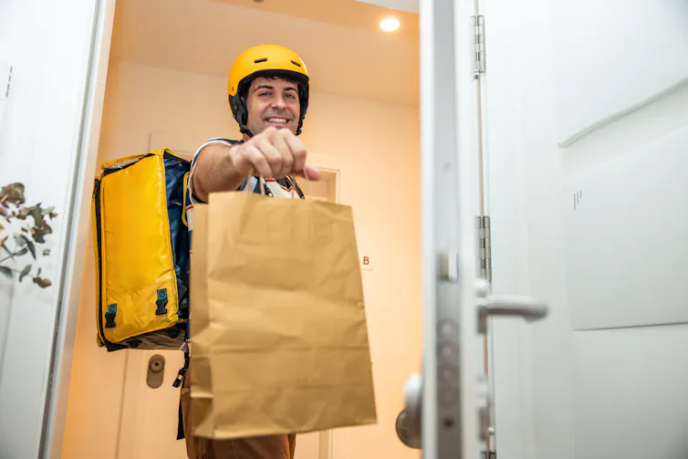 Man wearing a yellow helmet and carrying a thermal backpack delivering a takeout food order in a brown paper bag, standing at an open home door