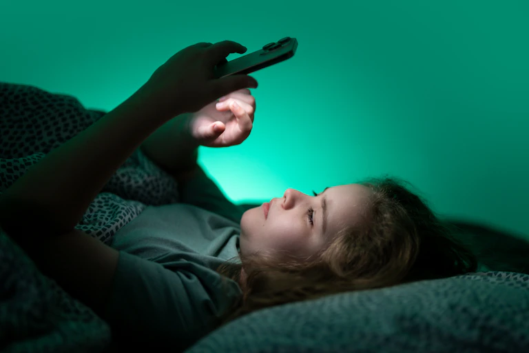 A teenager scrolling on a phone in bed at night.