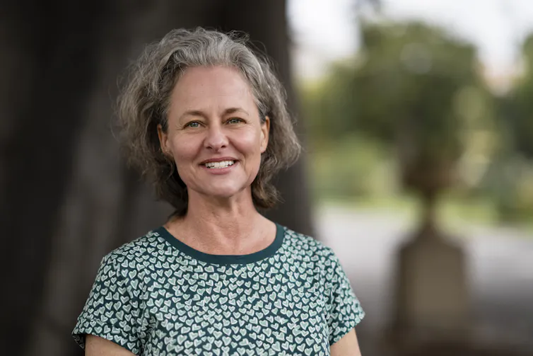 A grey-haired woman smiling outside, wearing a patterned green t-shirt.