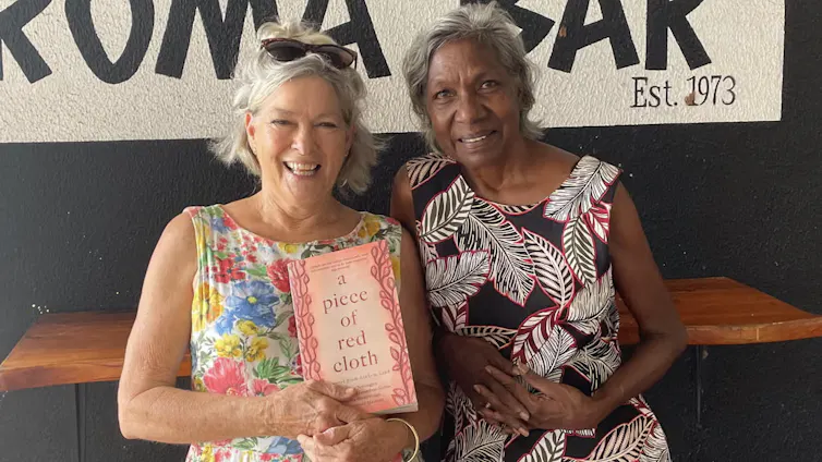 A white woman and a Yolŋu woman stand together, smiling and holding a book, wearing patterned shift dresses.