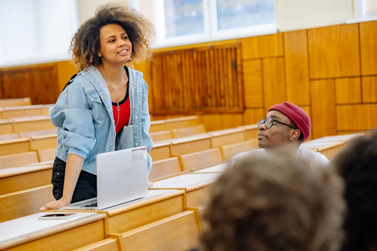 Young woman standing at a desk in a lecture theatre; young man looks on
